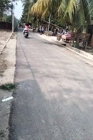 A paved road is visible with a grassy shoulder and some vegetation. A person is riding a motorcycle on the road. There are trees and structures on the left side of the road, with several people gathered. The scene appears to be outdoors during the daytime.