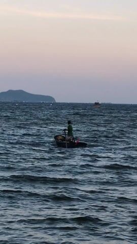 Here is a concise description of the video:

The video captures a single individual piloting a small, dark-colored boat across a choppy sea. The person, dressed in a green shirt, stands at the back of the boat, managing the boat's engine. In the distance, a dark, mountainous island is visible on the horizon, along with a faint glow of lights. The sky is an pale gradient of pink and gray from the horizon. The overall impression is one of solitude and the rhythm of the boat against the waves.