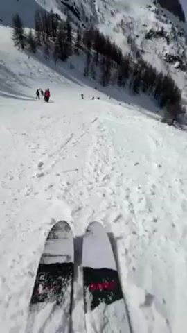 The video shows a first-person view of someone skiing down a snow-covered mountain slope. Other skiers are also visible on the slope. The skis are predominantly white with black and red graphics. There are trees on the sides of the slope and a mountain in the background. The sky is mostly clear.