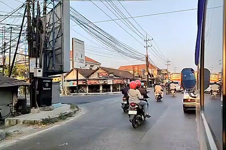 The video shows a street scene from the perspective of a vehicle, likely a bus, moving forward. The street is lined with buildings that appear to be shops or businesses, with awnings and signs. There are several motorcycles with riders on the road, some in front of and beside the camera's vehicle. Overhead, a complex network of electrical wires is visible against a clear sky. The sun appears to be low in the sky, suggesting it's either early morning or late afternoon. The overall atmosphere is that of a moderately busy street in what seems to be a town or city.