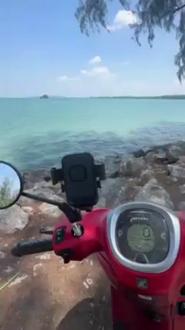 A still shot shows a red Honda Scoopy scooter parked on rocky ground next to a shoreline. The turquoise water of the sea extends to the horizon under a clear blue sky. A small island is visible in the distance. The scooter's handlebars and dashboard are in the foreground, featuring a phone mount. A selfie is reflected in the scooter's side mirror, revealing a person wearing a helmet. The scene is set in a sunny, beachside location.
