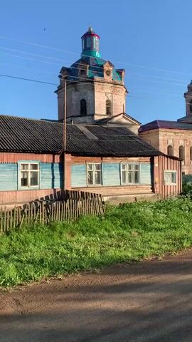 The video shows a side view of a rural church. In the foreground, there is a small wooden house with blue siding and a black roof. A wooden fence is in front of the house, and there is green grass and weeds around it. The church is in the background, made of brick with multiple domes. The main dome is larger and has a colorful, possibly green and red, roof. There is another, smaller dome on a bell tower. The sky is blue and clear, and the sun is shining. There are power lines visible in the upper part of the frame.