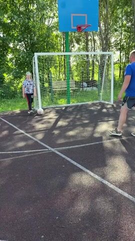 In an outdoor sports area with a dark red and black asphalt court, two individuals are playing soccer. The court has white markings and is bordered by a chain-link fence and trees. A soccer goal with a white net is visible, and above it, a blue basketball hoop with a red rim is mounted on a green pole. A young person with blonde hair, wearing a black and white patterned t-shirt and black pants with red stripes, is positioned on the left side of the soccer goal, near the ball. They are in the process of kicking the soccer ball towards the goal. To the right of the goal, a taller person, wearing a blue t-shirt and black shorts, stands facing the goal, seemingly in a defensive position. The lighting suggests it is daytime with sunlight casting shadows on the court.