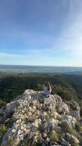 The video shows a woman sitting cross-legged on top of a rocky peak. She is wearing sunglasses, a light blue shirt, and blue pants, and has red shoes. The peak overlooks a vast landscape of green hills and plains under a blue sky with sparse clouds. The sun is visible in the upper right of the frame, casting a bright light. The video pans slightly to the right, shifting the focus from the woman to the surrounding landscape.