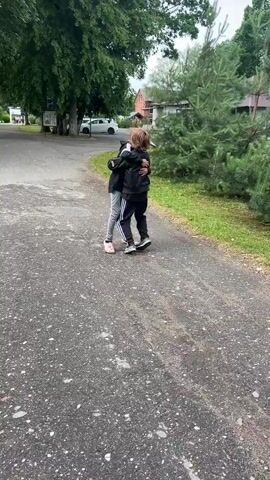 Two children, a girl and a boy, are walking on a paved path outdoors. The girl is wearing a black hooded sweatshirt with pink and white accents, grey pants, and pink Croc-like shoes. The boy is wearing a black jacket over a t-shirt, black pants with white stripes, and grey sneakers. They are initially hugging each other. The girl then puts on a grey baseball cap. They are both smiling and appear to be interacting with each other. The background shows trees, grass, a white car, and some buildings. The weather appears to be overcast.