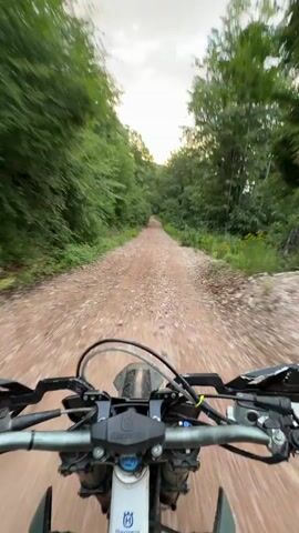 A first-person perspective video shows a dirt bike riding on a gravel road through a forest. The camera is mounted on the handlebars, providing a view of the road ahead and the surrounding trees. The sun is setting, casting a warm glow through the trees. The rider is wearing black gloves.