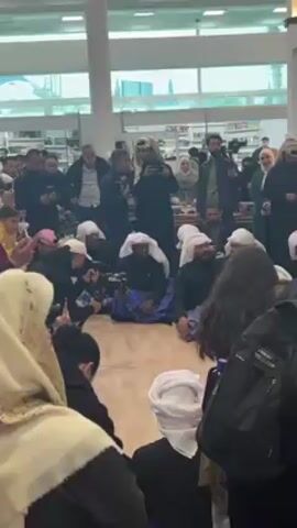 The video shows a group of men, some of whom are seated on the floor in a circle, wearing traditional Middle Eastern clothing. People are standing around the group, filming and photographing them. The background appears to be a public space like a library or a building with large windows and shelves. The video focuses on the men seated on the floor and the crowd surrounding them, suggesting a performance or cultural event.