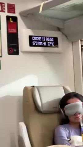 The camera is located inside a train. The train appears to be traveling at 173 kilometers per hour. A passenger is seated in a chair; they are wearing a sleep mask and headphones. On the wall is a digital display that reads "COCHE 11 08:50 173 KM/H 27°C." There is an alarm switch on the wall to the left of the digital display. Another passenger is seated next to the sleeping passenger and is holding a smartphone. A tray table is attached to the back of the sleeping passenger's chair; a brown paper bag is resting on it.