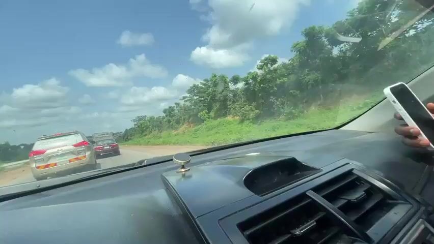 This media file captures a view from the dashboard of a car driving on a paved road in Aciol, Nigeria. The car is moving along a highway with a bright blue sky filled with fluffy white clouds overhead. Lush green vegetation lines the sides of the road, with trees and shrubs creating a vibrant natural backdrop.

In front of the car, several other vehicles are visible. The closest is a light-colored SUV, possibly a Toyota, with tinted windows and what appears to be signage on its rear windshield and doors. Following this SUV is a darker sedan. Further down the road, another dark-colored car is also in motion.

The road itself appears to be a multi-lane highway with a clear white line marking the lanes. The surrounding landscape suggests a rural or semi-rural setting, with occasional glimpses of more distant trees and foliage. The bright daylight and clear skies indicate it is daytime and the weather is fair. There are no visible people within the frame of the car's interior, other than the implied driver and passenger whose perspective the video offers.

The overall impression is of a journey on a sunny day through a green and somewhat populated area of Nigeria. The presence of multiple vehicles suggests ongoing traffic and the use of this road for transportation.