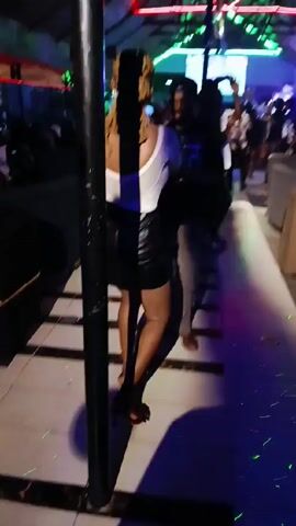 The video depicts people dancing in a nightclub setting. The main focus is on a woman in a white top and black skirt, moving to the music. The nightclub has a tiled floor with a geometric pattern. There are other people visible, some dancing and some standing around. The lighting is colorful and typical of a nightclub. The environment is dimly lit with bright, colorful accent lighting. The overall atmosphere suggests a social gathering with dancing and music.