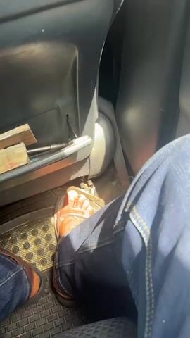 This is a first-person perspective video from inside a vehicle. The camera is focused on the legs and feet of the person recording. They are wearing blue jeans and brown dress shoes. Their right foot is pressing down on what appears to be the accelerator pedal of the vehicle, indicated by the shoe's position on the pedal and the slight flexion of the ankle. The left foot is also visible, resting on the floor mat. The interior of the car is dimly lit, with sunlight filtering in from an unseen window. The dashboard and seat are partially visible. There is a black rubber floor mat with a raised, patterned texture. A small compartment or tray is visible to the left, containing what appears to be a pen and some papers. The video is shaky, suggesting movement, consistent with being inside a moving vehicle.