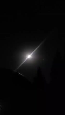 The video shows a nighttime scene with a bright moon visible in a dark sky. A sharp beam of light emanates from the moon, extending diagonally across the frame. Silhouettes of trees are visible on the right side of the image, and a curved object, possibly the roof of a vehicle, is in the lower left corner. The scene appears to be outdoors.