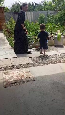 A woman in a long dark floral dress stands on a paved area, looking to her left. A young boy in a blue t-shirt and shorts stands facing away from the camera, holding a red plastic cup and a blue bucket, near a water faucet and a drain. He is stooped over and appears to be interacting with the water. Behind them is a garden with raised beds of plants, a metal fence, and trees.
