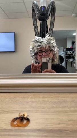 A woman with glasses and short blonde hair wrapped in foil and cotton is holding a smartphone and taking a selfie in the mirror. She is smiling. Above her head is a black and silver hair dryer. Two earrings are on the table in the foreground. Behind the woman in the mirror is a washing machine, a shelf stocked with bottles, and a television on the wall.