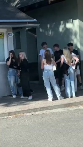 A group of about nine teenagers are standing on a sidewalk next to a building. The teenagers appear to be conversing and socializing. Some are looking at their phones. The building is light green with a darker green roof extension. There is a white box with signage on the side of the building. The sidewalk is made of gray pavers, and a street is visible in the foreground. The scene is brightly lit, suggesting it is daytime.