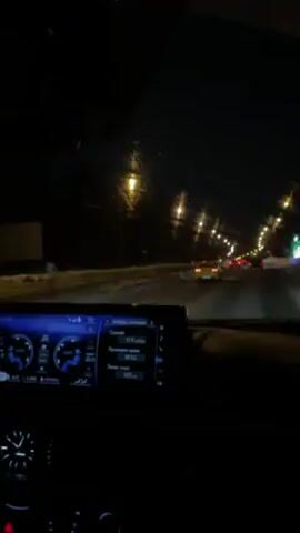 The video shows a view from inside a car driving at night on a road with moderate traffic. The road appears to be covered in snow or slush. Streetlights illuminate the scene. The car's dashboard is visible, displaying information on a digital screen. The windshield wipers are in motion. The overall impression is of a nighttime drive in winter conditions.