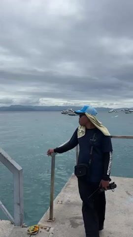 The video shows a man in a blue hat and a dark blue shirt standing on a pier. He is looking out at the water, which has several boats in the distance and mountains on the horizon. The sky is overcast. The man has a towel around his neck and is carrying a walkie-talkie. Another person walks by with a cell phone in their hand. There is a small offering on the pier by the steps.