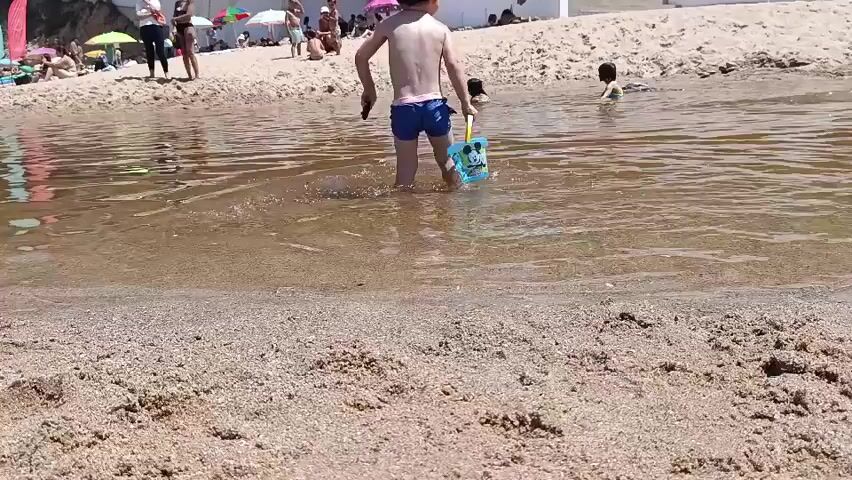 This video captures a sunny day at a beach with a river meeting the sea. The foreground is dominated by sandy beach with ripples from the water. In the middle ground, a young boy in blue swim trunks walks through shallow water, carrying a blue bucket with a Mickey Mouse design and a red toy shovel. The water is murky brown, with ripples indicating movement.

In the background, the beach is populated with many people enjoying the day. Several individuals are sitting or standing near the water's edge. Further back, a variety of beach umbrellas provide shade, suggesting a warm, sunny day. Some people are lying on the sand, and a few are in the water. The setting appears to be a popular beach destination. The overall mood seems relaxed and cheerful, characteristic of a summer beach outing. The time of day is likely midday, given the bright sunlight and shadows. There is no visible text in the video.