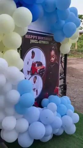 The video shows a decorated outdoor space, presumably for a child's birthday party. There's a large sign that says "Happy Birthday Joel-Ethan" with photos of the child. The area is decorated with blue, yellow, and white balloons arranged in arches and clusters. There's also a life-size banner with a photo of a young boy in a blue shirt and white pants. We can see a woman in a green dress passing by. There are also decorations in the shapes of jeeps. The setting appears to be a lawn or garden area outside a building or house, with a black wrought-iron fence partially visible in the background. There is loud celebratory music playing.
