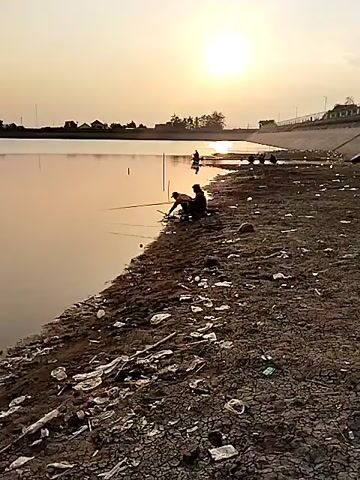 A wide shot of a body of water reflecting a sunset. Several people are fishing on the bank of the water. A road or path runs along the top of a concrete embankment. The embankment is littered with trash. The video pans to the right, showing more of the embankment and a building in the distance.
