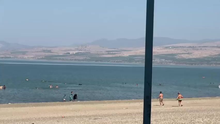 The video shows a beach scene with a large body of water and distant hills. The sky is clear and hazy. On the sandy beach, a vertical dark pole is partially visible on the right side. Several people are in the water, swimming and playing. Near the shore, a few individuals are standing on the beach, including a man and a woman with a hula hoop. Further left, a group of people are near the water's edge, with some appearing to be children. The water is calm and appears to be a lake. The environment suggests a warm, sunny day suitable for recreational activities at the beach.