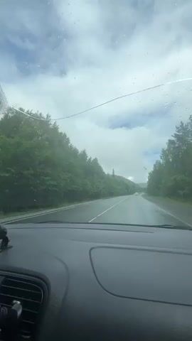 The video shows a view from inside a car driving on a wet road lined with trees on both sides. The sky is cloudy with patches of blue visible. There are raindrops on the windshield. The road appears to be a two-lane highway. The car's dashboard is visible in the foreground. The overall atmosphere is overcast and damp.