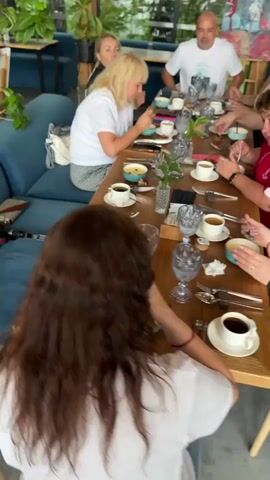 A group of eight people are seated around a long wooden table, which is set with plates, cups, and cutlery. The people are of various ages and genders. They appear to be dining or having coffee. The room has large windows with a view of greenery and trees. There are also many plants inside the restaurant. The camera is moving, giving a panning view of the scene.