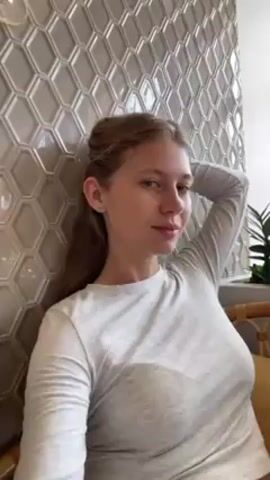 A light-skinned woman with blonde hair is the main subject of the video, which appears to be taken from a close-up, slightly low angle. She's wearing a light grey long-sleeved shirt. The woman is leaning against a wall covered in geometric light grey tiles and has one arm raised behind her head. Her hair is partially pulled back from her face. She looks into the camera, smiles briefly, and touches her temple with her fingers. In the background, there's a glimpse of a plant in a wicker basket and what may be a light fixture.