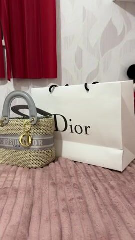A still shot shows a small, woven straw handbag with grey leather handles and a grey band across its front with "CHRISTIAN DIOR PARIS" written in white lettering. A gold "D" charm hangs from the handle. Next to the handbag is a white Dior shopping bag with black handles and the word "Dior" printed in black. In the background, red curtains hang to the left, and a textured wallpaper covers the wall behind the bags. A small Mickey Mouse plush toy peeks over the top of the Dior shopping bag. The items are placed on a pink, textured surface that resembles a blanket.