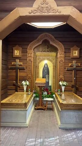 The video shows the interior of a wooden room, likely a chapel or a church. In the center, there is an icon of a bearded man in religious attire holding a scroll, framed by intricate wooden carvings. In front of the icon is a wooden lectern with an open book. Flanking the icon are two large, ornate wooden sarcophagi or reliquaries, each topped with glass and adorned with floral arrangements. Two wooden crosses are mounted on the wooden walls on either side of the central icon. The walls and ceiling are made of dark wood, and the floor appears to be paved with brick or stone. The lighting is dim, with a soft glow emanating from above the icon and from within the sarcophagi. There are no people visible in the video.