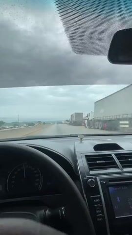 The media file shows a view from inside a car, driving on a highway in Kwali, Nigeria. The scene is primarily of the road ahead, with traffic visible.

**Subjects:**
*   **Objects:** A car interior (steering wheel, dashboard, center console with infotainment system and air vents), vehicles on the highway (including trucks and cars), roadside structures that appear to be market stalls or shops, and a large container truck with "MAERSK" written on it.
*   **Scene/Setting:** A highway in Nigeria, with a wide road and a shoulder area. The surroundings include a slightly hilly landscape with scattered trees and vegetation. There are also what appear to be small buildings and informal market stalls lining the side of the road. The sky is cloudy, suggesting an overcast day.

**Activity or Event:**
The primary activity is driving on a highway. There appears to be some congestion or slow-moving traffic, as several vehicles are visible in the distance and on the side of the road.

**Notable Details:**
*   **Time of Day:** Based on the lighting and the cloudy sky, it appears to be daytime, likely late morning or afternoon.
*   **Weather:** The sky is heavily overcast with clouds, indicating potentially rainy or gloomy weather.
*   **Location Cues:** The presence of Maersk container trucks, the style of roadside structures, and the general landscape are consistent with locations in Nigeria. The dashboard clock shows "12:49".
*   **Emotions:** The perspective from the car suggests a journey in progress. The cloudy weather and traffic might imply a less than ideal driving experience, but no strong emotions are directly conveyed.

**Visible Text:**
*   "MAERSK" is clearly visible on a large container truck.
*   The car's dashboard displays speed readings (up to 220), "PWR", "VOL", "AUDIO", "SEEK", and "TRACK" on the infotainment system, and the time "12:49".
