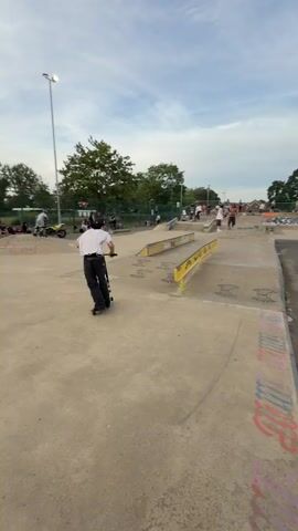 The video depicts a man riding a scooter at a skatepark. He is wearing a white t-shirt, black pants, and a black baseball cap. The man attempts to ride the scooter on a low yellow rail. He loses his balance and falls backwards off the rail, nearly face planting. There are other people at the skatepark riding bikes and scooters, watching. The skatepark is made of concrete and features various ramps and rails. There is graffiti on the skatepark's surfaces, and the sky is partly cloudy.