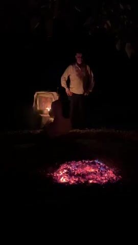 The video displays a low-light outdoor scene at night. In the foreground, a pile of glowing embers, radiating red and blue light, rests on the ground. Behind the embers, a person is kneeling on the ground facing away from the camera, illuminated by a soft light source. To the left of the kneeling person, a small, illuminated altar-like structure is visible, draped with fabric. Standing behind the kneeling person and to the right is another individual, a man, facing the camera and appearing to observe the scene. The overall environment is dark, suggesting it is nighttime, with faint outlines of foliage visible in the upper portion of the frame. The scene appears to be set in a natural, possibly wooded, outdoor area.