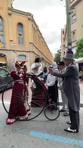 The video shows a group of people in vintage attire posing with a vintage bicycle on what appears to be a street or pedestrian area.  The bicycle has a large front wheel and a small rear wheel. A woman in a maroon dress and hat stands beside the bicycle, seemingly adjusting something near the handlebars.  Around her are four other individuals.  One woman wears a white dress with a large collar and a feathered hat. Another woman is in a black dress, white shirt, and a smaller hat.  Two men are dressed in suits and hats, one in a light grey suit and the other in a darker grey one. The man in the dark grey suit appears to be taking a photo with a smartphone. The background shows a building with numerous arched windows and doors, typical of older European architecture, with signage in a foreign language. A black van is visible on the left side of the frame. The scene seems to be part of a historical reenactment or celebration.