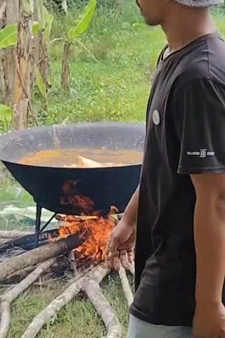 A person in a black shirt and light-colored pants stands next to a large black cauldron set over an open fire of burning wood. The cauldron is filled with a liquid that appears to be simmering. The fire is built on the ground, and the surrounding area is covered in green grass and some debris. The setting appears to be outdoors, possibly in a rural area.