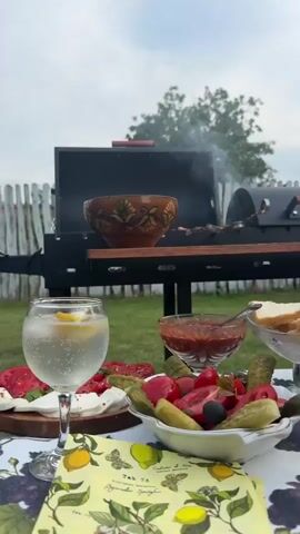 The video shows a close-up view of a table set up outdoors for a meal, with a grill in the background. On the table, there's a glass of clear liquid with ice and a lemon wedge, a bowl of red sauce, a bowl of chopped vegetables including tomatoes and pickles, a plate of sliced bread, and a small bowl of olives. A beer mug and a bottle of beer are also visible. The grill has food cooking on skewers, and smoke is rising. The background shows a wooden fence and some greenery, indicating a backyard setting. The camera focuses on the food and drinks, creating an inviting scene.