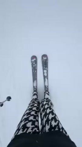 A person is skiing down a snowy slope. They are wearing black and white patterned ski pants. The skis are gray with red accents. The person is holding ski poles. The snow is white and appears soft.