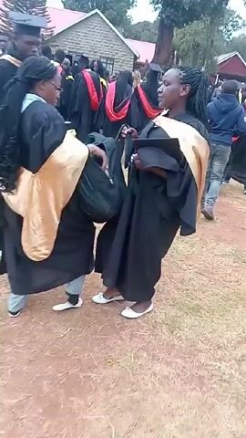 People wearing black graduation gowns and mortarboards are walking on a path. Some of the gowns have colored stoles in red and yellow. The path is dirt and grass. A person in casual clothing is walking among the graduates. A red banner with white lettering is visible in the background.