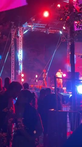 The video captures a lively outdoor concert at night.  A man is on stage, holding a microphone and singing, illuminated by red and blue stage lights. He is wearing a dark t-shirt and shorts. In front of the stage, a crowd of people are visible, silhouetted against the lights, some are facing the stage, others are interacting amongst themselves. The stage is set up with lighting rigs and speakers. Trees are visible behind the stage, suggesting an outdoor venue. The overall atmosphere appears energetic and festive.