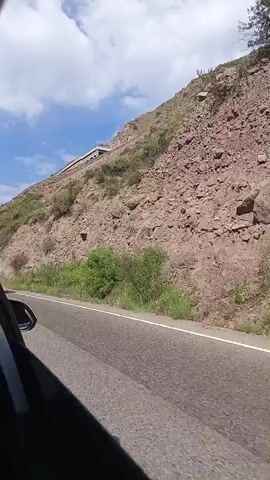 The video shows a view from a car window as it travels along a paved road. To the right of the road is a steep, rocky mountainside covered in sparse vegetation. The sky above is blue with scattered white clouds. A white SUV is seen in the distance driving in the opposite direction. The camera is angled to capture the passing scenery.