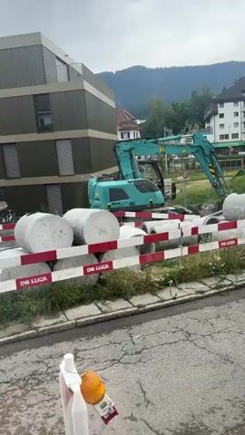 The video shows an excavator in a construction area. It is a turquoise wheeled excavator with a person in the driver's seat. The excavator is next to a modern building with dark brown siding and many windows. In front of the excavator are several large concrete pipes or culverts, and a red and white striped barrier. The ground appears to be a paved road or construction site, with some cracking and puddles of water. In the background, there are other buildings and a hillside with trees. The sky is overcast. The excavator appears to be maneuvering around the pipes and barriers, possibly preparing to move them or begin other construction work. There are a few other vehicles visible in the distance.