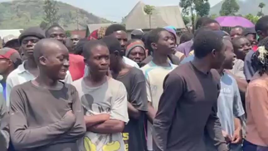 A crowd of mostly young, Black men are standing in a line in an outdoor area. The men are of varying heights, and they are dressed in casual clothing. Some of the men are smiling and talking, while others are looking around and listening. There is a mountain visible in the background.