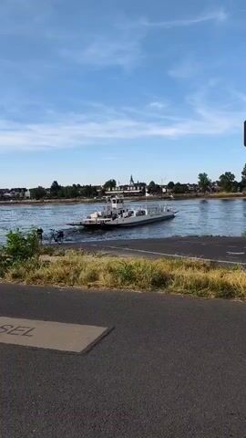 A wide shot shows a river with a ferryboat in the middle. The ferry is a flat-bottomed vessel with a superstructure in the center. It appears to be moving from left to right across the frame. On the far bank of the river, there are buildings, trees, and a spire-like structure. In the foreground, there is a paved path and some vegetation. The sky is blue with scattered clouds. Two people are visible on bicycles on the left side of the frame near the riverbank. The word DIESEL is imprinted on a square concrete slab in the foreground.