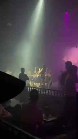 The video shows a group of young women dancing and drinking at a club. They are standing on a raised platform, separated from the rest of the crowd by a metal railing. The women are dressed in revealing outfits, mostly in black. There are two men in black suits standing guard, one on each side of the platform. The club is dimly lit with purple and white lighting, and there is a lot of smoke in the air. The music is loud and energetic.