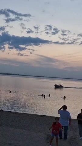 The video captures a serene evening scene at a body of water, likely a lake or a large river, as indicated by the Kokshetau, Kazakhstan location context. The sky is painted with the soft hues of dusk, transitioning from a pale blue at the top to warmer pinks and oranges closer to the horizon, with scattered clouds adding texture.

In the foreground, a sandy or gravelly shore is visible. On the shore, there are a few people. A young girl in a red shirt and dark shorts is bent down, appearing to be engaged in some activity on the ground. Behind her, a man in a light blue shirt and dark pants stands looking out towards the water. Another person is partially visible on the far right, also looking towards the water.

The water itself is calm, reflecting the colors of the sky. Several people are swimming or wading in the water, creating ripples. Further out on the water, a small motorboat with at least two occupants is moving, leaving a wake behind it. The overall atmosphere is peaceful and relaxed, suggesting a leisurely evening by the water. The time of day is clearly sunset or twilight, given the sky's coloration. There is no visible text in the video.