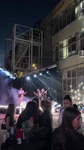 This is a vertical, handheld video taken at night. In the background, a band is performing on a stage lit by bright white spotlights and some colored lights, with smoke effects. There are at least three musicians visible on stage, one playing guitar and singing, another playing guitar, and a third in the center. In the foreground, several people, mostly young women, are standing and sitting at tables, facing the stage. The environment appears to be an outdoor or semi-outdoor venue with industrial-looking buildings and scaffolding in the background. The lighting is dramatic, with strong contrasts between light and shadow. The overall mood seems to be a lively concert or performance.