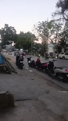 The video shows a street scene at dusk. There are several motorcycles parked along the side of the road, and a few cars are driving on the road. There are trees on either side of the street, and buildings are visible in the background. The sky is a light gray color, indicating that the sun is setting or has just set. The overall impression is one of a typical urban or suburban street at the end of the day.
