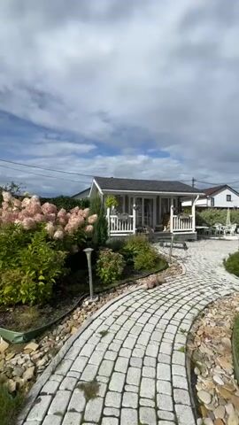 The video shows a close-up view of a garden with blooming pink hydrangeas in the foreground. In the background, a small white shed with a porch is visible, along with a paved walkway made of light-colored stones. The sky is partly cloudy, with white clouds against a blue background. The camera pans slightly, moving closer to the hydrangea bush and then slightly away, revealing more of the shed and the garden path.