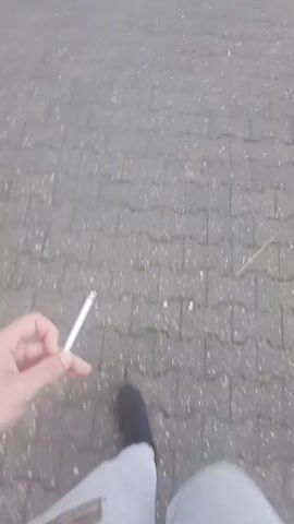 A person is walking down a paved sidewalk on a foggy morning. The person is wearing light gray sweatpants and black sneakers with a pink accent. They are holding a lit cigarette in their right hand. The sidewalk is bordered by a building on the left and parked cars on the right. A light pole is visible on the right side of the sidewalk. As the person walks, the fog dissipates slightly, revealing more of the buildings and cars.