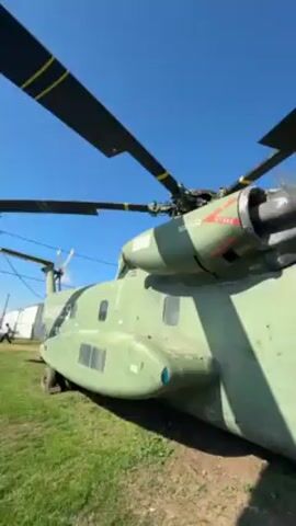 The video shows a large, green military helicopter on a grassy area under a clear blue sky. The helicopter has large rotor blades and the words "MARINES" and "RESCUE" are visible on its exterior. A man wearing a bright green shirt and jeans is standing inside the open doorway of the helicopter. A set of metal steps leads into the helicopter. The camera pans slightly, showing the helicopter's cockpit window and the inscription "COL. DICK WILSON MAG 'MAD DOG'."